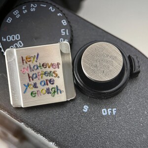 May include: A square silver-toned metal camera hot shoe cover featuring the colorful engraved text "hey! whatever happens, you are enough." and a circular silver-toned metal camera hot shoe cover with the same text engraved in black.
