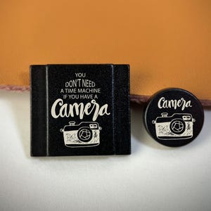 Könnte beinhalten: Zwei schwarze Accessoires zum Thema Kamera. Eines ist quadratisch und trägt den Text "YOU DON'T NEED A TIME MACHINE IF YOU HAVE A CAMERA" mit einer Kamera-Illustration. Das andere ist rund, mit dem Wort "Camera" und einer Kamera-Illustration.