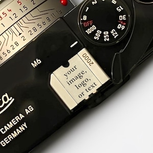 May include: A black Leica M6 camera featuring a silver metal hot shoe cover engraved with the text 'your image, logo, or text'. The camera body displays the Leica logo and 'LEICA CAMERA AG GERMANY' text, with a shutter speed dial and lens visible.