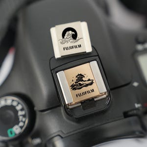 Puede incluir: Primer plano de una cámara Fujifilm negra con una cubierta de zapata plateada y dorada. La cubierta presenta un diseño de olas negras con pájaros y el texto "FUJIFILM". Una segunda cubierta está encima con un diseño similar.