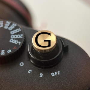 Op de afbeelding: Close-up van een zwarte camera met een goudkleurige draaiknop met de letter "G". De knop bevindt zich boven de bedieningselementen van de camera, inclusief "OFF". De instelknop van de camera is links zichtbaar.