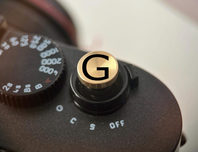 Op de afbeelding: Close-up van een zwarte camera met een goudkleurige draaiknop met de letter "G". De knop bevindt zich boven de bedieningselementen van de camera, inclusief "OFF". De instelknop van de camera is links zichtbaar.