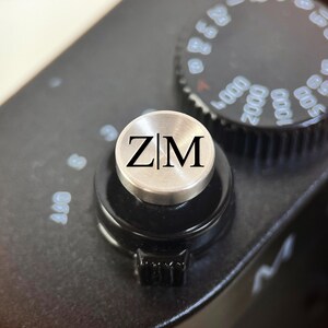 Op de afbeelding: Close-up van een zwarte camerabody met een zilverkleurige knop. De knop heeft de zwarte letters "ZM". Een zwarte draaiknop met witte cijfers is zichtbaar op de achtergrond.