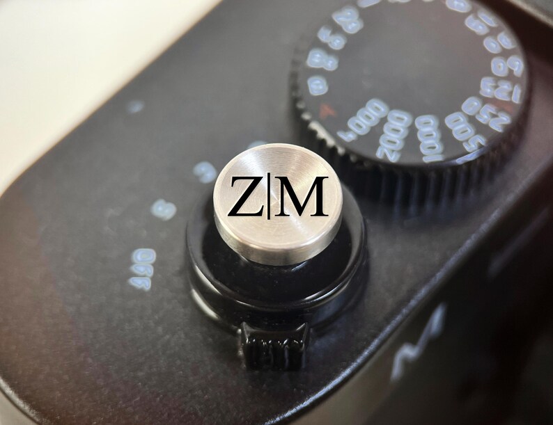 Op de afbeelding: Close-up van een zwarte camerabody met een zilverkleurige knop. De knop heeft de zwarte letters "ZM". Een zwarte draaiknop met witte cijfers is zichtbaar op de achtergrond.