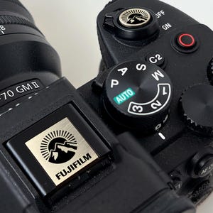 Puede incluir: Primer plano de una cámara Fujifilm negra con un logotipo plateado y dorado. La lente indica 2.8/24-70 GMI. La cámara tiene varios diales y botones, incluido un dial de modo automático y un interruptor de encendido/apagado. El logotipo presenta un diseño de montaña y sol.