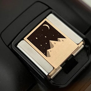 May include: A close-up of a gold-colored flash attachment for a camera. The attachment has a black and white design of a mountain range with a crescent moon and stars.