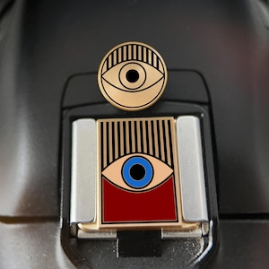 May include: A circular gold-tone enamel pin featuring a black eye graphic with vertical lines above it, positioned above a rectangular gold-tone enamel pin displaying a blue eye graphic with vertical black lines above and a solid dark red base.