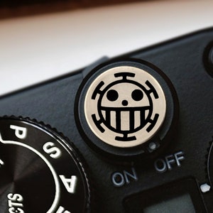 May include: A black camera with a silver knob featuring a black and white skull and crossbones symbol. The camera has a black and silver dial with the words "SCENE", "AUTO", "P", "S", and "A" printed on it. The camera also has a black and silver button with the word "ON" printed on it and a black and silver button with the word "OFF" printed on it.