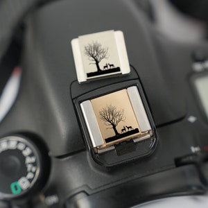 May include: A black camera with a silver flash attachment. The flash attachment has a small, rectangular, gold-colored insert with a silhouette of a tree and two deer. The camera has a dial on the side with numbers and a small green rectangle.