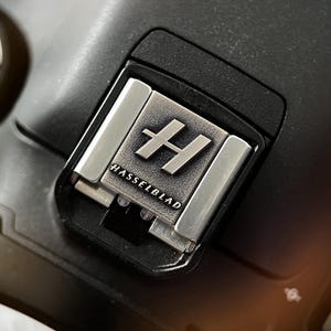 Op de afbeelding: Close-up van een zwarte camera met een zilveren Hasselblad-logo. Het logo is een gestileerde "H" in een vierkant, met "HASSELBLAD" eronder gedrukt. De flitsschoen van de camera is zichtbaar, met zilveren metalen accenten.