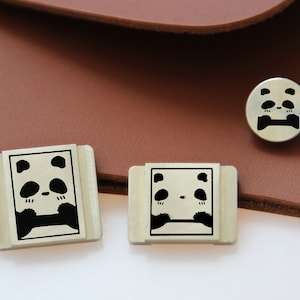 May include: Three silver metal panda bear charms. The charms are in different sizes. The largest charm is rectangular and has a panda bear face with closed eyes. The medium charm is also rectangular and has a panda bear face with closed eyes. The smallest charm is round and has a panda bear face with closed eyes.