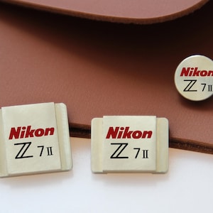 May include: Three Nikon Z 7II camera accessories. Two rectangular accessories and one circular accessory, all with the Nikon logo in red and black. The accessories are on a light brown surface.