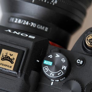 Puede incluir: Primer plano de una cámara Sony negra con una lente. La cámara presenta un botón dorado con un diseño floral y un logotipo Fujifilm dorado. La lente está etiquetada FE 28/24-70 GM.