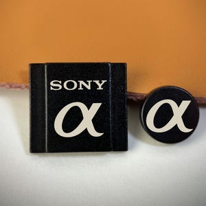 May include: A black rectangular camera hot shoe cover featuring the white text SONY and the Greek alpha symbol, alongside a small black circular button cover displaying the same white Greek alpha symbol on its flat surface.