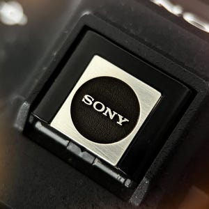 Puede incluir: Primer plano de un componente negro y plateado con el nombre de la marca "SONY" en letras blancas. El logotipo está centrado en un cuadrado plateado, rodeado por un marco negro. El fondo es oscuro y borroso.