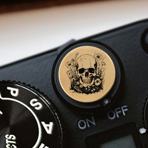 May include: A close-up of a black camera with a gold-colored dial featuring a black skull design surrounded by flowers. The camera's settings dial is visible, with the words "ON" and "OFF" on the dial.