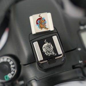 May include: A black camera with two flash shoe covers. One cover features a colorful tiger design with a red sun and blue waves. The other cover displays a black and white tiger illustration.