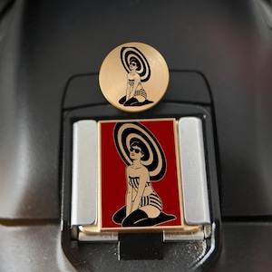 May include: A circular gold-toned metal pin and a rectangular gold-toned metal pin, both featuring a black and white illustration of a person wearing a large striped hat, sunglasses, a striped bikini, and black stockings against a deep red background.