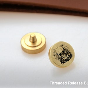 May include: A gold-colored threaded release button with a skull and flowers design. The button is a small, round, metal piece with a threaded hole in the center. The design is engraved on the top of the button.
