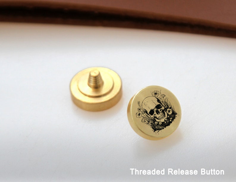 May include: A gold-colored threaded release button with a skull and flowers design. The button is a small, round, metal piece with a threaded hole in the center. The design is engraved on the top of the button.
