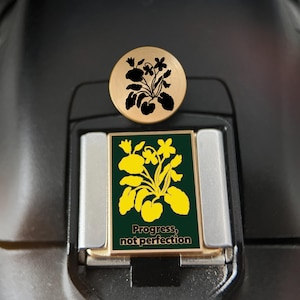 Può includere: Una spilla rotonda color oro con un'illustrazione floreale nera e una spilla rettangolare color oro con sfondo verde scuro, un'illustrazione floreale gialla e il testo "Progress, not perfection" stampato in un carattere scuro e grassetto.