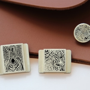 May include: Three silver metal stamp designs featuring black and white zebra patterns. The stamps are rectangular and have rounded corners. The stamps are on a brown leather surface.