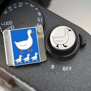 May include: A rectangular metal camera hot shoe cover featuring a blue background with a large white goose and three smaller white goslings, alongside a circular silver shutter button cover engraved with a single black line drawing of a goose.