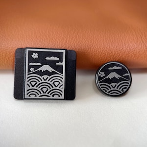 Könnte beinhalten: Eine rechteckige schwarze Anstecknadel und eine runde schwarze Anstecknadel, beide mit einer silberfarbenen Gravur eines Berggipfels, stilisierten Wellen, Wolken und einer kleinen Blume. Die Anstecknadeln haben eine matte Oberfläche.