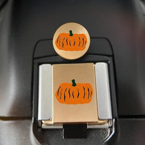 Puede incluir: Una tapa para zapata de cámara de metal en tono dorado con una parte superior circular y una base rectangular, ambas decoradas con un gráfico de calabaza naranja brillante con un tallo verde oscuro y detalles en negro para la zapata.