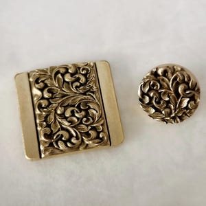 以下が含まれることがあります： 複雑な花の模様が施された、金色の金属製のボタン2つ。大きいボタンは正方形、小さいボタンは丸形です。