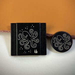 Puede incluir: Un sujetador de agujas rectangular negro y uno circular negro a juego, ambos con una ilustración de líneas blancas de un pulpo rodeado de pequeñas burbujas y un pequeño icono de planta en la pieza rectangular.