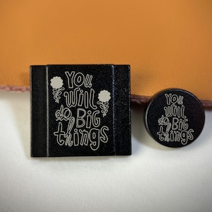 Puede incluir: Una etiqueta rectangular negra y un botón circular negro, ambos con el texto blanco grabado "You will do Big things" acompañado de pequeñas ilustraciones de flores. Los artículos tienen un acabado liso y letras claras y legibles.