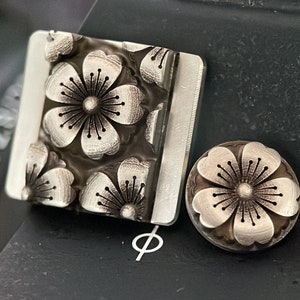 May include: Two silver metal flower-shaped buttons with a detailed design. The larger button has multiple flowers, while the smaller button has a single flower.