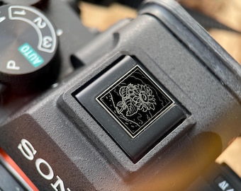 May include: Close-up of a black camera body with a silver and black floral design. The design is a square-shaped emblem with a detailed outline of a sunflower. The camera has a mode dial and the brand name "SONY" is visible.