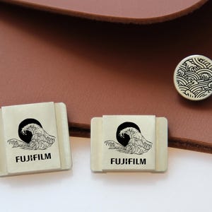 Puede incluir: Accesorios Fujifilm color crema con un diseño de olas negras y el logotipo de Fujifilm. También se ve un botón redondo con un patrón de olas. Los artículos están colocados sobre un fondo de cuero marrón.