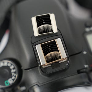 以下が含まれることがあります： 黒いデジタル一眼レフカメラのクローズアップ。上部に2つの銀色のフレームの長方形アクセサリーが付いています。アクセサリーは、曲線のある白黒のデザインです。左側には緑色のマーキングが付いたダイヤルが見えます。