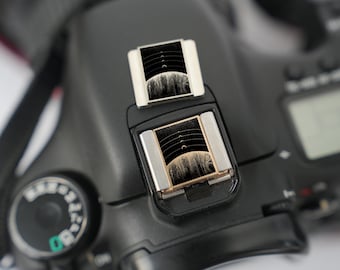May include: Close-up of a black DSLR camera with two silver-framed, rectangular accessories on top. The accessories have a black and white design with curved lines. A dial with green markings is visible on the left.