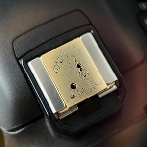 Puede incluir: Una cubierta de zapata de flash de cámara de metal dorado con un diseño grabado de un astronauta, un cohete y estrellas. La cubierta está unida a un cuerpo de cámara negro.