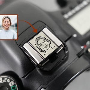 Puede incluir: Una cámara negra con una montura de flash plateada. La montura del flash presenta un dibujo lineal en blanco y negro de una mujer sonriente. Una imagen insertada muestra una foto en color de la misma mujer.