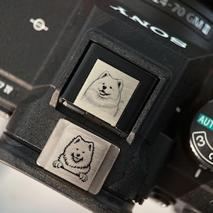 May include: A black camera with a silver hot shoe cover featuring an engraved image of a white dog. The camera has a dial with the word "AUTO" and numbers 1, 2, and 3.