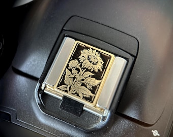 May include: Close-up of a camera's hot shoe cover. The cover is rectangular with a gold and black floral design. The design features a sunflower and other flowers. The cover is framed by silver metal.