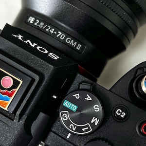 Könnte beinhalten: Nahaufnahme einer schwarzen Sony-Kamera mit Objektiv. Die Kamera hat ein Moduswahlrad mit Einstellungen wie 'AUTO' und 'M'. Dekorative Anstecknadeln mit farbenfrohen Landschaftsdesigns sind sichtbar.