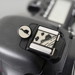 Puede incluir: Primer plano de una cámara negra con una montura de flash plateada. Se ve un botón redondo plateado con un gráfico de pato negro. La montura del flash tiene un gráfico en blanco y negro de un pato y un paisaje.