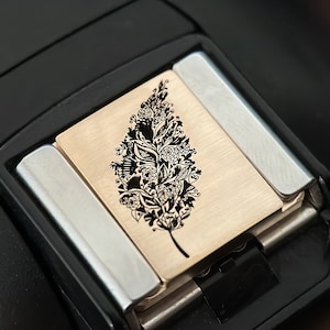 Pode incluir: Um close-up de um isqueiro preto e dourado com um design floral decorativo. O isqueiro apresenta uma placa retangular dourada com um padrão floral preto detalhado. O isqueiro é envolto em uma moldura preta com detalhes prateados.