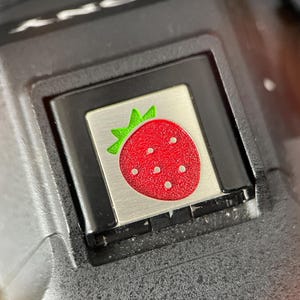 May include: Close-up of a camera button featuring a strawberry design. The button is silver with a red strawberry and green leaves. The camera body is black.