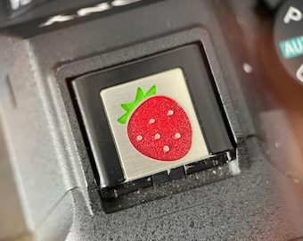 May include: Close-up of a camera button featuring a strawberry design. The button is silver with a red strawberry and green leaves. The camera body is black.