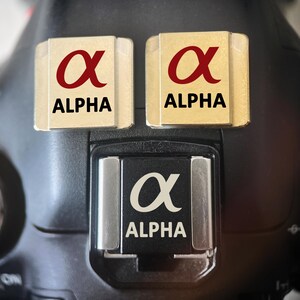 Pode incluir: Close-up de um corpo de câmera preto com três detalhes do logotipo "Alpha". Duas placas retangulares douradas com o símbolo "Alpha" vermelho e a palavra "ALPHA" em texto preto estão no topo. Um terceiro logotipo "Alpha" está na montagem do flash da câmera.
