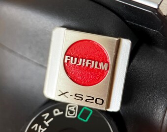May include: A silver and red Fujifilm X-S20 camera badge. The badge has a silver frame with the word "FUJIFILM" in white letters on a red circle. The text "X-S20" is printed in black below the circle. The badge is on top of a black camera.