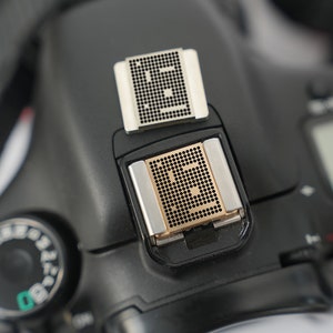 May include: Close-up of a black camera with two small, square, metal flash units attached to the top. The flash units have a grid pattern.