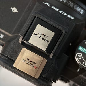 May include: Close-up of a black camera body with a silver Fujifilm X-T30 II label and a gold Fujifilm X100V label. The camera has a lens attached and a dial with AUTO, 3, 2, 1, P, and A settings.